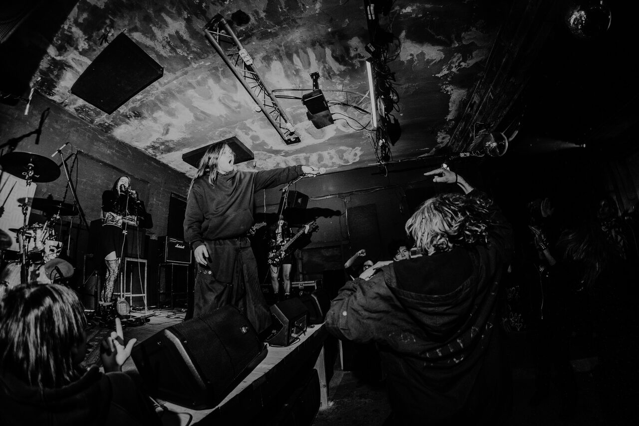 Арендовать PVZ CLUB (ПВЗ Клуб). Чёрно-белое фото со сценическим музыкальным выступлением в тёмном помещении. На сцене микрофоны, барабаны, колонки, освещение сверху. Потолок с эффектом разрушенности, стены без декора, присутствуют люди.