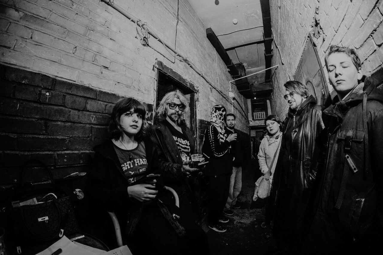 Арендовать PVZ CLUB (ПВЗ Клуб). узкий коридор с кирпичными стенами и низким потолком; группа людей, некоторые сидят на стульях, другие стоят; видна проводка и вентиляционная труба на потолке; черно-белое изображение.