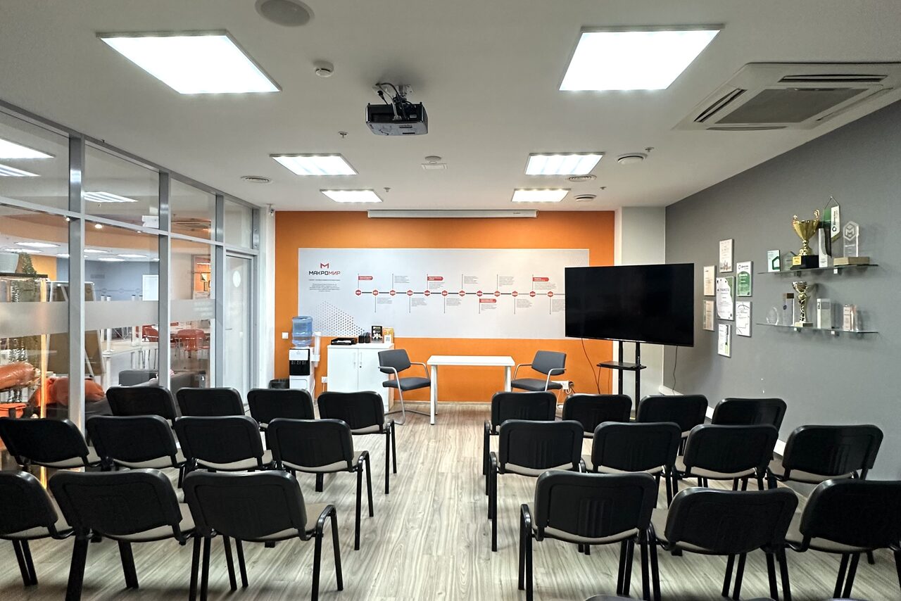 Арендовать Конференц-зал. Конференц-зал с рядами черных стульев, белыми и оранжевыми стенами. На переднем плане телевизор на штативе, белый стол, стеклянные двери. Светлое деревянное покрытие пола, потолочные светильники, проектор. На стенах полки с наградами и плакатами.
