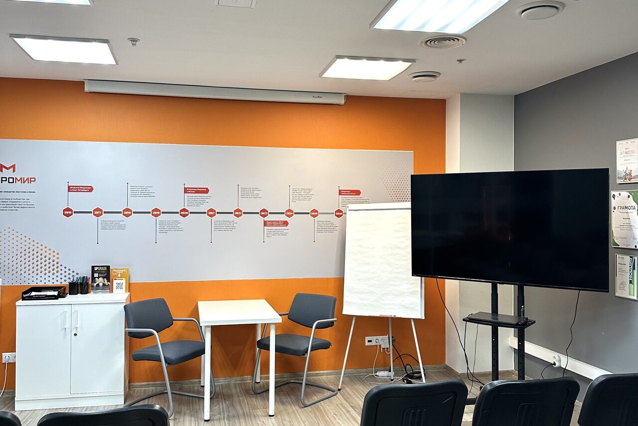 Арендовать Конференц-зал. Совещательная комната с белой и оранжевой стеной, настенным плакатом. Круглый стол, два стула, флипчарт и большой экран на стойке. Белый шкаф, потолочный светильник, кондиционеры, деревянный пол. Несколько плакатов на стене.