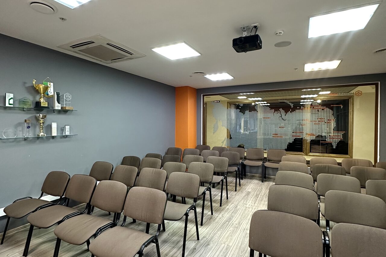 Арендовать Конференц-зал. Комната с расположенными рядами стульев. На серой стене полки с наградами. Потолочные светильники, проектор, кондиционер. Стеклянная перегородка с картой, деревянный пол. Цвета: серый, оранжевый, коричневый.