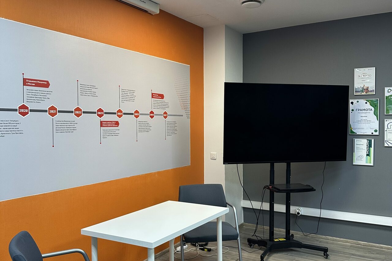 Арендовать Конференц-зал. Современное офисное пространство с серыми стенами и акцентной оранжевой стеной. На оранжевой стене инфографика. В комнате белый стол, серые стулья, большой телевизор на стойке. Справа на стене сертификаты и документы. Пол серый.