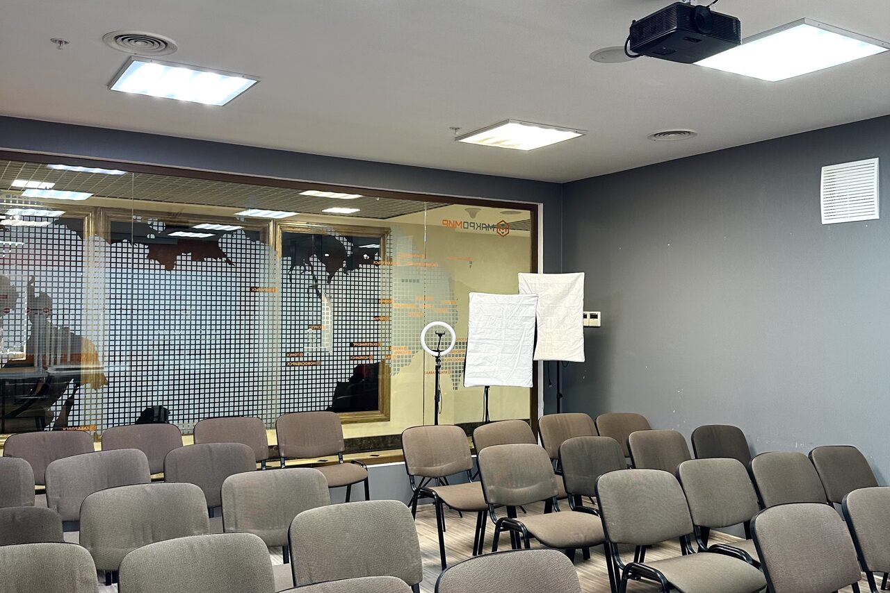 Арендовать Конференц-зал. Конференц-зал с серыми стенами, множеством серых стульев, стеклянной перегородкой с нанесенным рисунком на заднем плане. На потолке светильники и проектор. Рядом с перегородкой световое оборудование и кольцевая лампа.