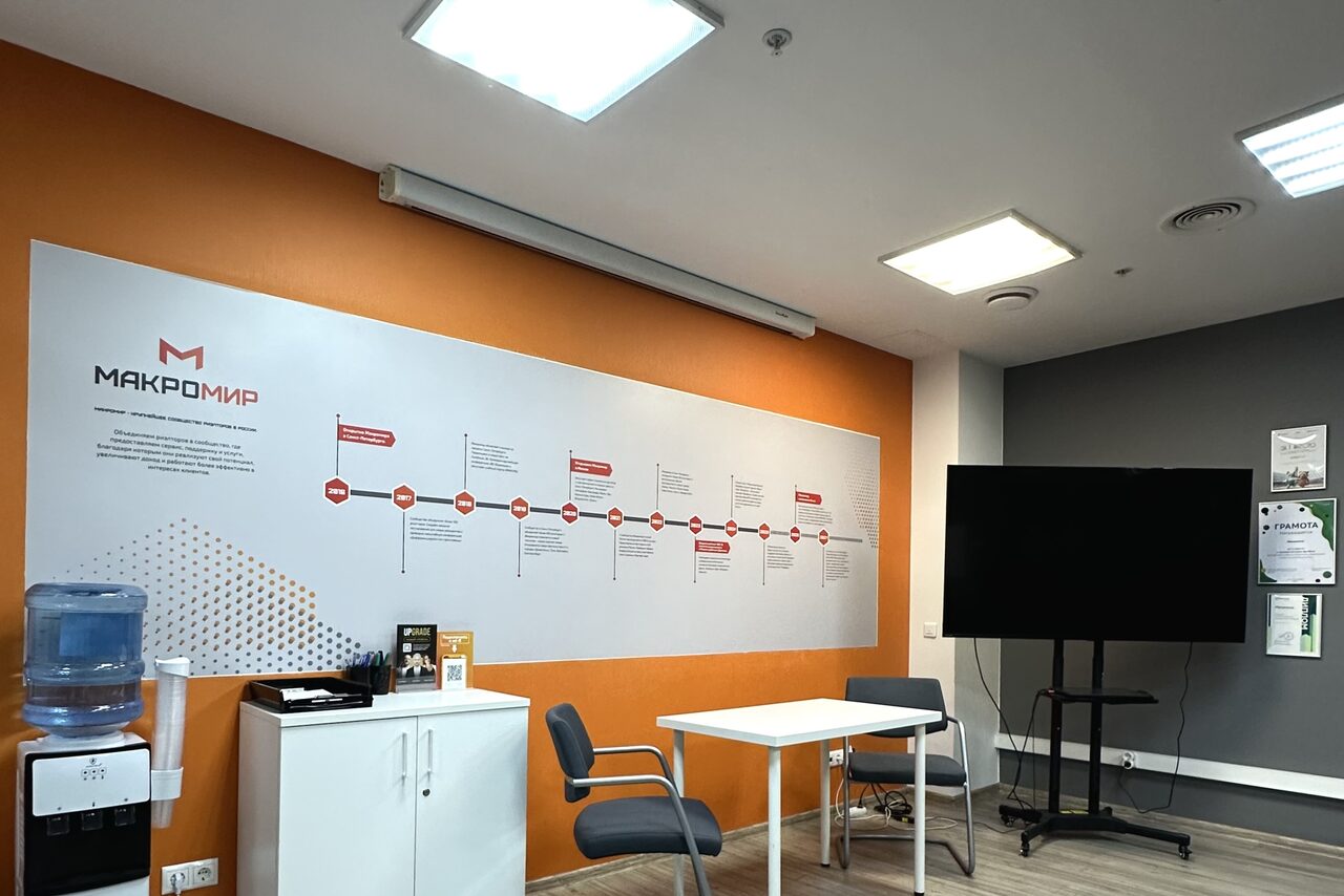 Арендовать Конференц-зал. Современный офис. Оранжевая и серая стены, на оранжевой стене информационная панель. Белый шкаф с кулером, два стула, стол. Большой экран на черной стойке. Яркое потолочное освещение. На стене несколько напечатанных материалов.