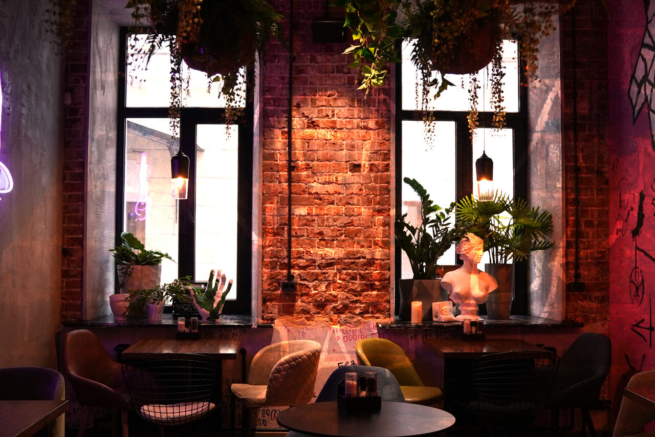 Арендовать Винный бар “Дело не в вине”. Лофт-пространство с кирпичными стенами. Два окна с чёрными рамами, декоративные растения на подоконниках и бюст. Низкие чёрные столы, разноцветные стулья. Подвесное освещение, неоновые элементы, стены с граффити. Атмосферное освещение.