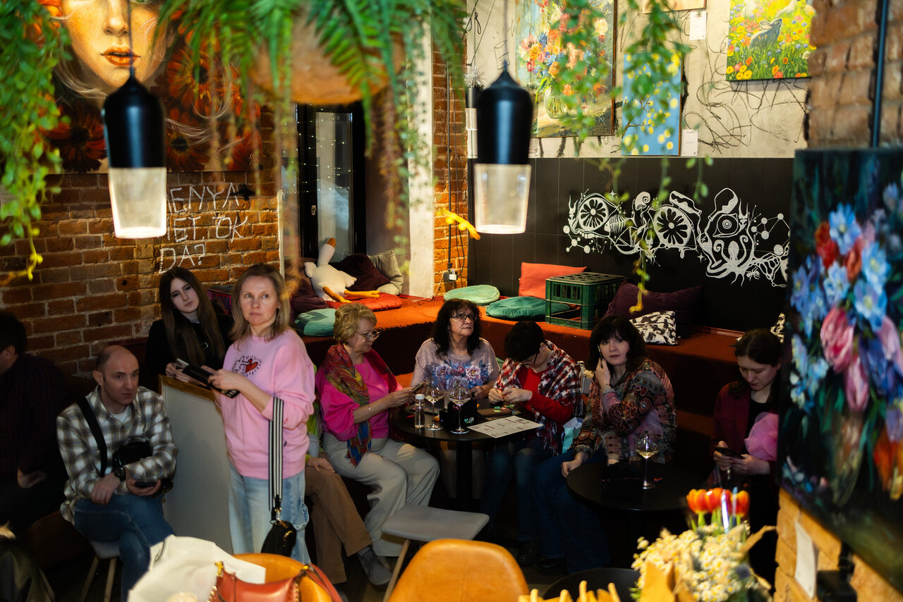 Арендовать Винный бар “Дело не в вине”. Кафе или лаунж пространство с кирпичными стенами и настенными рисунками. Мягкие сиденья с подушками, столики, люди сидят и стоят, растения свисают с потолка. Освещение с подвесными лампами, картины на стенах.