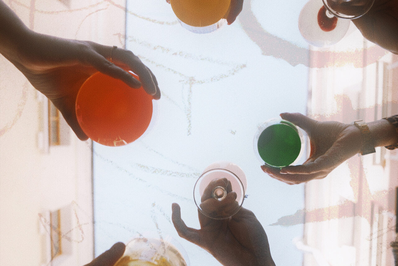 Арендовать Винный бар “Дело не в вине”. Руки людей держат разноцветные стеклянные бокалы на фоне светлого неба и размытых очертаний зданий.