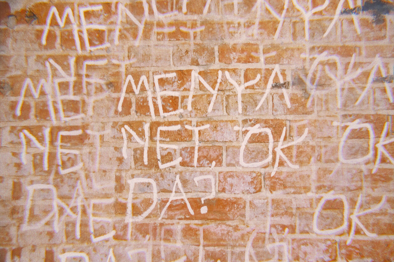 Арендовать Винный бар “Дело не в вине”. Кирпичная стена с надписями белой краской, включая слова "МЕНЯ", "НЕТ", "OK", "ГДЕ?". Текст выполнен крупными небрежными буквами, фон состоит из стандартной красно-коричневой кирпичной кладки.