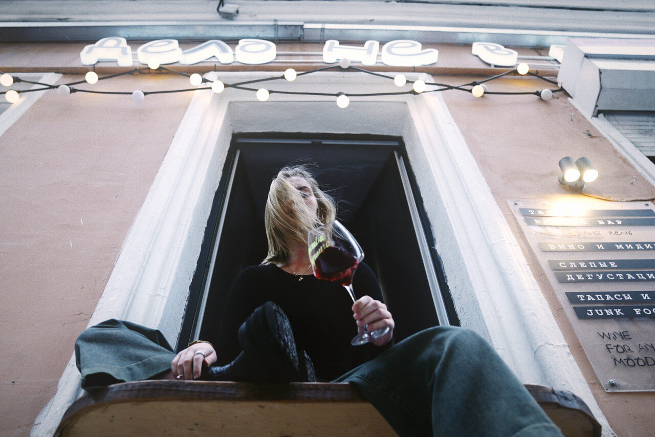 Арендовать Винный бар “Дело не в вине”. Наружный фасад здания с неоновой надписью и гирляндой лампочек. Женщина сидит на подоконнике, держа бокал вина. Справа от окна на стене прикреплена доска с текстом. Фасад окрашен в светло-оранжевый цвет.