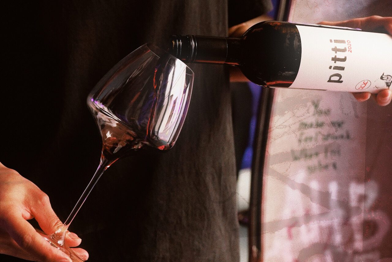 Арендовать Винный бар “Дело не в вине”. Человек наливает красное вино в бокал из бутылки с белой этикеткой. Тёмный фон с размытыми деталями.