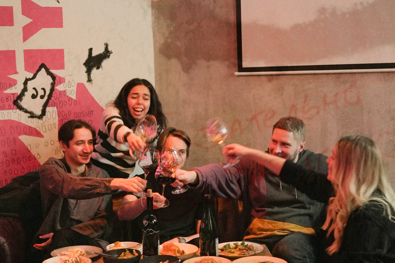 Арендовать Винный бар “Дело не в вине”. Изображение показывает людей за столом в помещении с бетонными стенами и граффити. На столе тарелки с едой и бутылки. Люди держат бокалы. На фоне виден проектор или экран. Освещение тёплое.