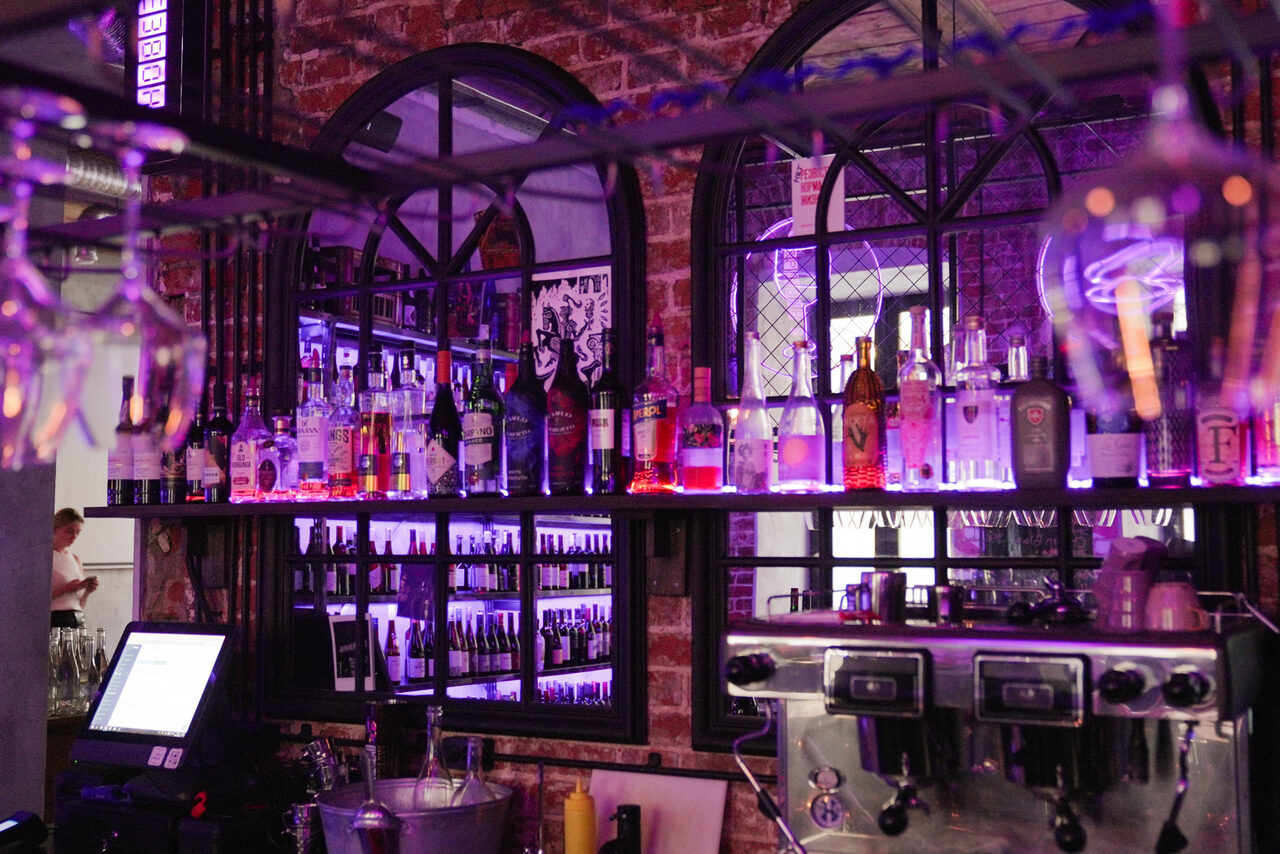 Арендовать Винный бар “Дело не в вине”. Барная стойка с кирпичной стеной, стеклянные полки с алкогольными бутылками, фиолетовая подсветка, зеркала с арочными рамами, кофеварка, место кассира с монитором, подвешенные бокалы, металлические элементы интерьера.