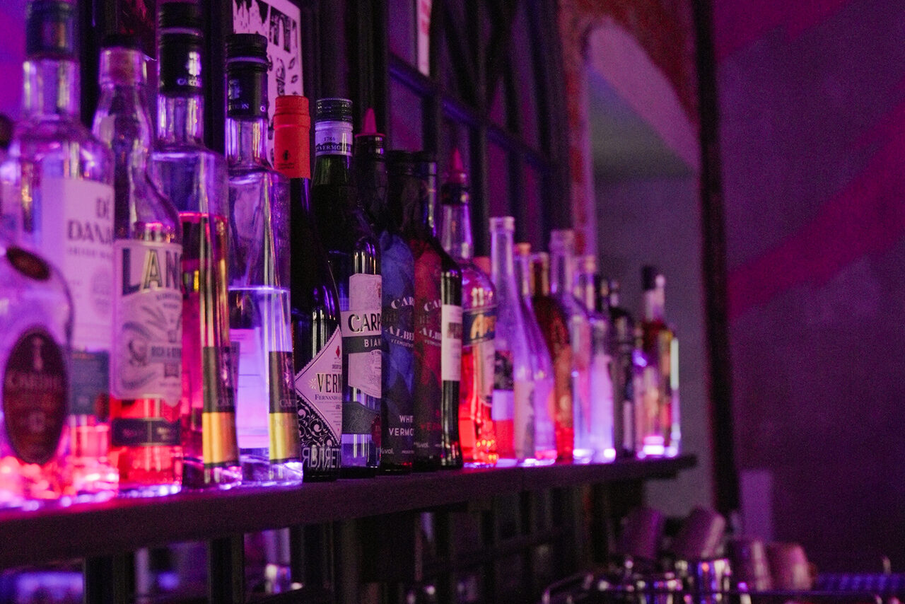 Арендовать Винный бар “Дело не в вине”. Полка с бутылками спиртных напитков в баре, подсветка фиолетового цвета, темная рама оконной конструкции на фоне, различные формы бутылок, яркие этикетки, стеклянная посуда, часть фиолетовой стены.