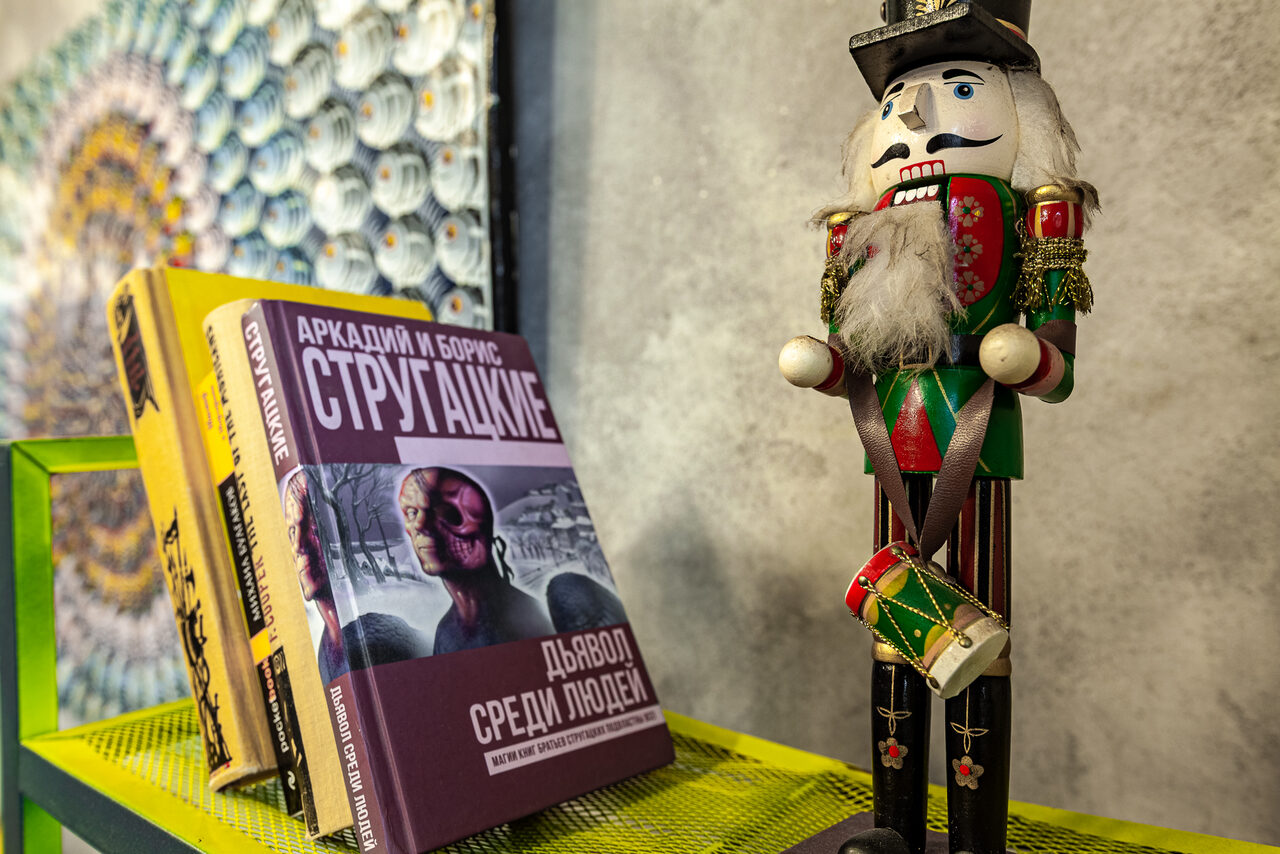 Арендовать Лофт где можно все. Книги на полке и деревянная фигурка щелкунчика. Стена из серого бетона, на фоне разноцветный абстрактный рисунок. Полка из жёлтого металла с ячейками. Книги стоят вертикально, одна из них лежит обложкой вверх.