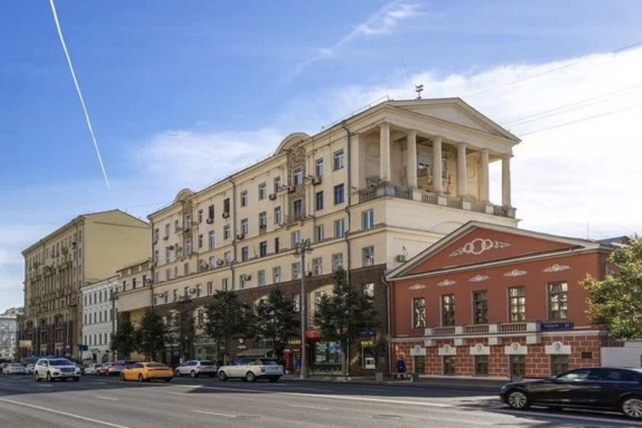 Арендовать Лофт где можно все. Улица с автомобильным движением. Слева многоэтажное здание с колоннами и балконами светлых тонов. Справа — одноэтажное здание красного цвета с белыми декоративными элементами. Ясное небо с самолетным следом.