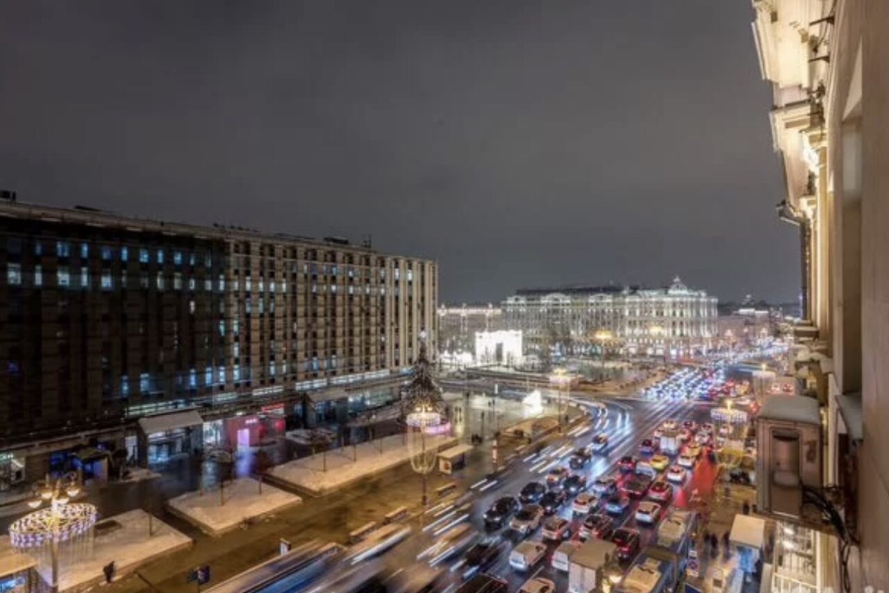 Арендовать Лофт где можно все. Вид из окна на оживлённую городскую улицу ночью. Движение автомобилей, свет фар. Высотные здания по обеим сторонам улицы. Освещение от уличных фонарей. Пасмурное небо.