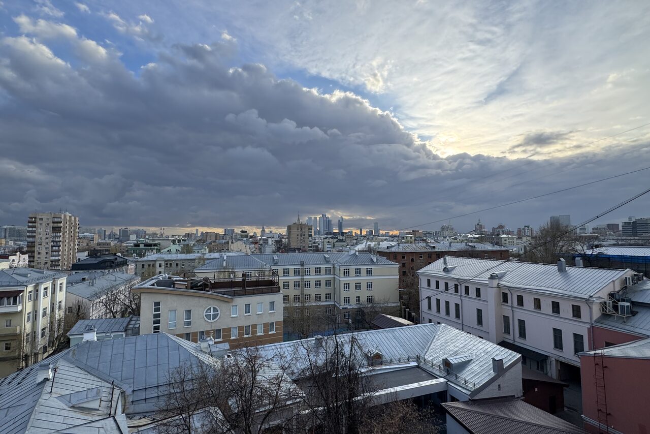 Арендовать Лофт где можно все. Вид на городские крыши с высоты; облачное небо с просветами на закате; различные многоэтажные здания с белыми, розовыми и бежевыми фасадами; множество окон; металлические крыши; видны bare деревья.