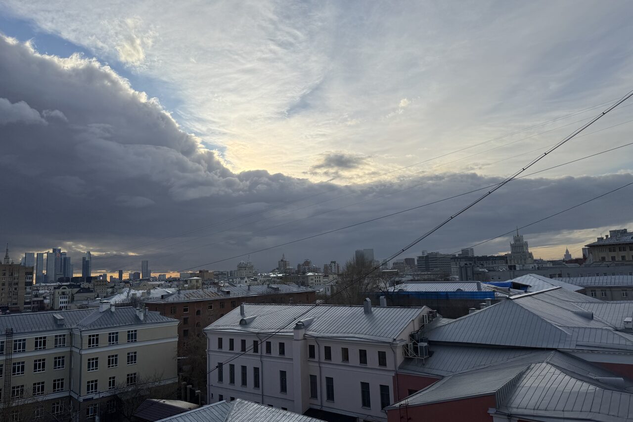 Арендовать Лофт где можно все. Вид с высоты на городской пейзаж с облачным небом, несколькими зданиями с серыми крышами и виднеющимися вдалеке небоскрёбами. Яркое освещение рассвета или заката.