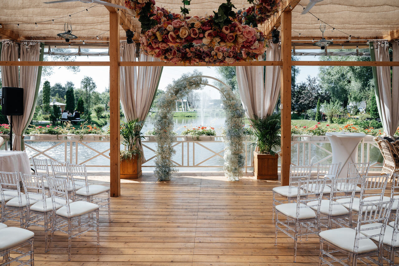 Арендовать Веранда Картмазовские пруды. Открытая терраса с деревянным полом и тканевым потолком. Панорамные окна с видом на водоём и зелень. Арка из цветов и растения в горшках. Ряды прозрачных стульев с белыми сидениями. Белые скатерти на высоких столах. Сверху висят гирлянды из лампочек.