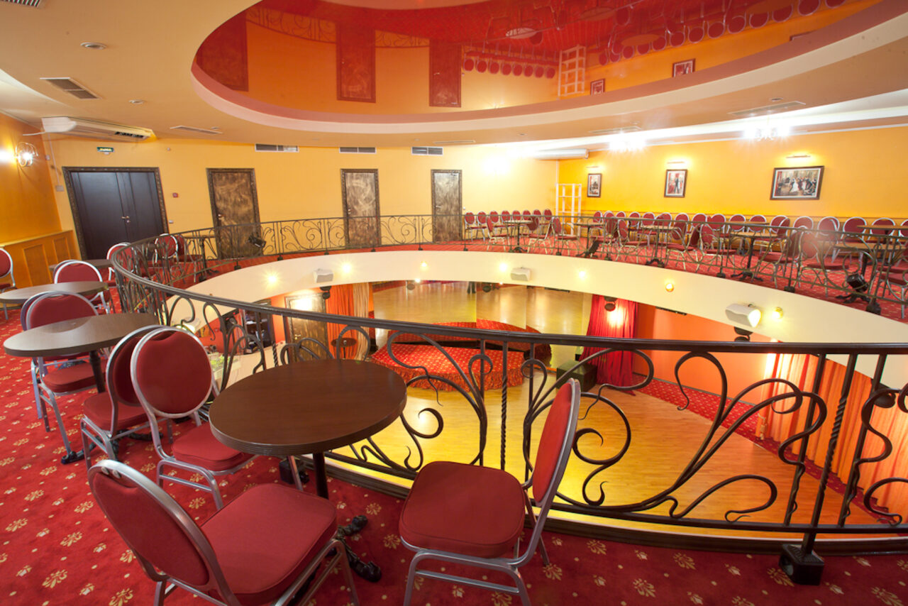 Арендовать Jenavi CLUB(Дженави Клуб). Круглый зал с балконом и перилами, множество красных стульев и столы. Освещение точечное, на стенах картины. Пол и ковровое покрытие красные с узором. Стены желтые, двери темные.