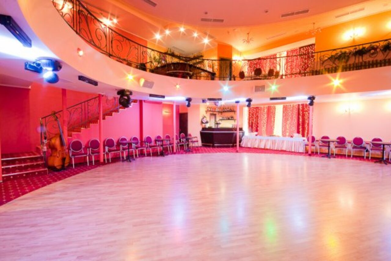 Арендовать Jenavi CLUB(Дженави Клуб). Пространство с круглой планировкой, паркетный пол, по периметру розовые стулья, два уровня с перилами и лестницей слева, барная стойка, яркое освещение с цветными лампами, стены розового и оранжевого цвета, задрапированные окна.