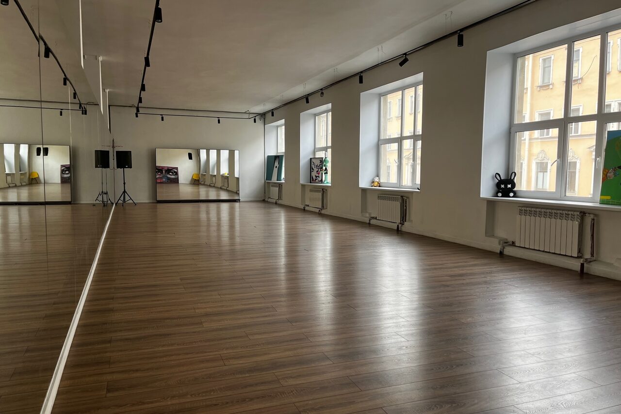 Арендовать The Flow Space(Пространство Поток). Просторное помещение с деревянным полом и большими окнами, длинные зеркала на одной стене, белые стены, потолок с черными светильниками, радиаторы под окнами, два черных акустических динамика.