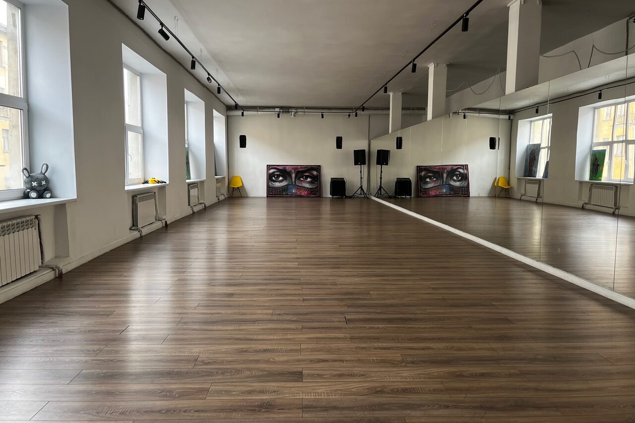 Арендовать The Flow Space(Пространство Поток). Просторная комната с деревянным полом и большими окнами вдоль стены. На одной стене — зеркало во всю длину. На дальнем конце — звукосистема и настенные панели с изображением глаз. Помещение хорошо освещено, светлые стены, черные светильники на потолке.