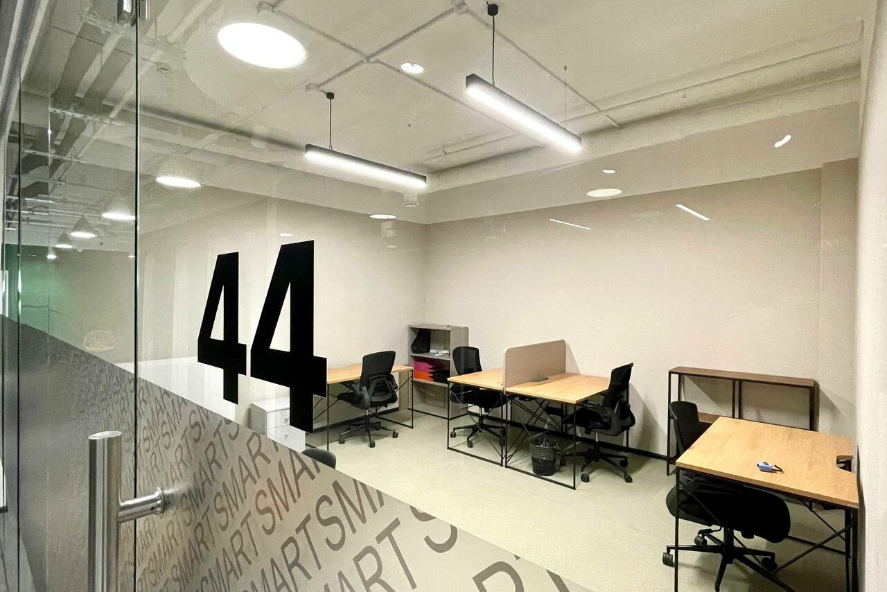 Арендовать Smart Coworking(Умный Коворкинг). Офисное помещение с бежевыми стенами. Четыре стола и кресла, полки для документов. Стеклянная дверь с номером "44". Потолочные светильники. Ламинат серого цвета на полу. На стекле повторяющийся текст "SMART".