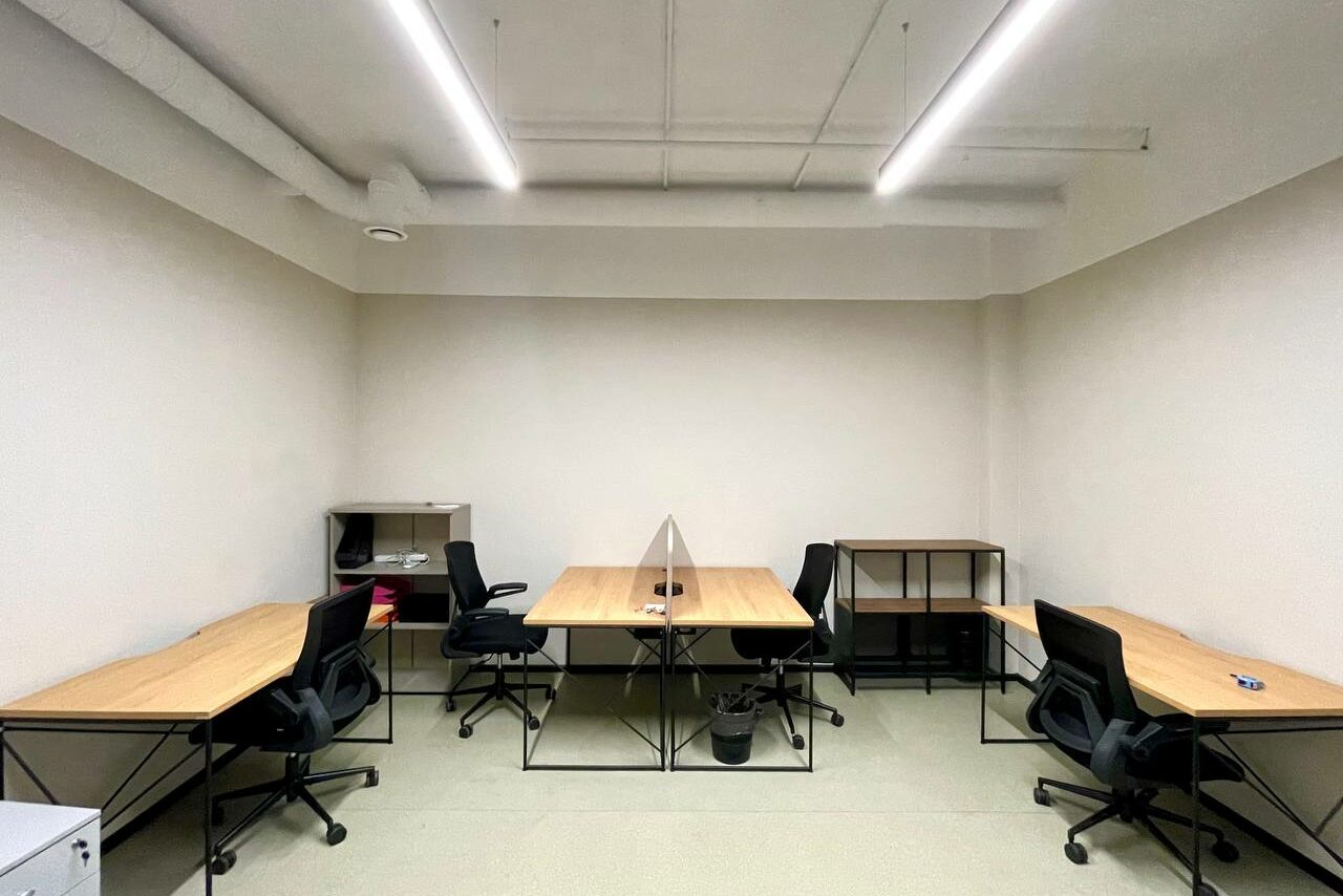 Арендовать Smart Coworking(Умный Коворкинг). Офисное помещение с пятью рабочими местами. Планировка линейная с расположением столов вдоль стен. Каждый стол оснащён чёрным офисным креслом. Светлые стены и пол, нейтральное освещение, параллельные потолочные светильники.