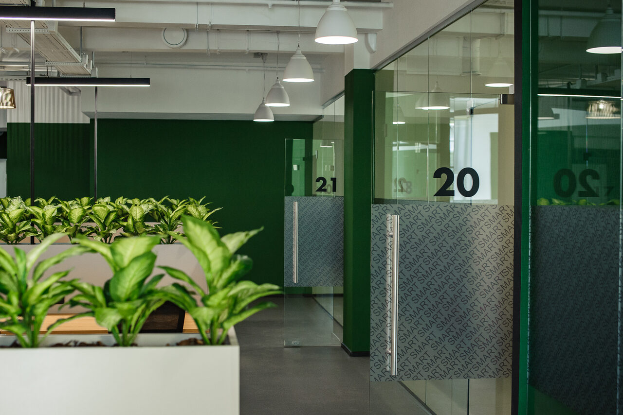 Арендовать Smart Coworking(Умный Коворкинг). Офисное пространство с прозрачными стеклянными перегородками и дверями, пронумерованными цифрами 20. Вдоль стен расположены кашпо с зелеными растениями. Потолочные светильники белого цвета. Доминируют зеленые и нейтральные тона.