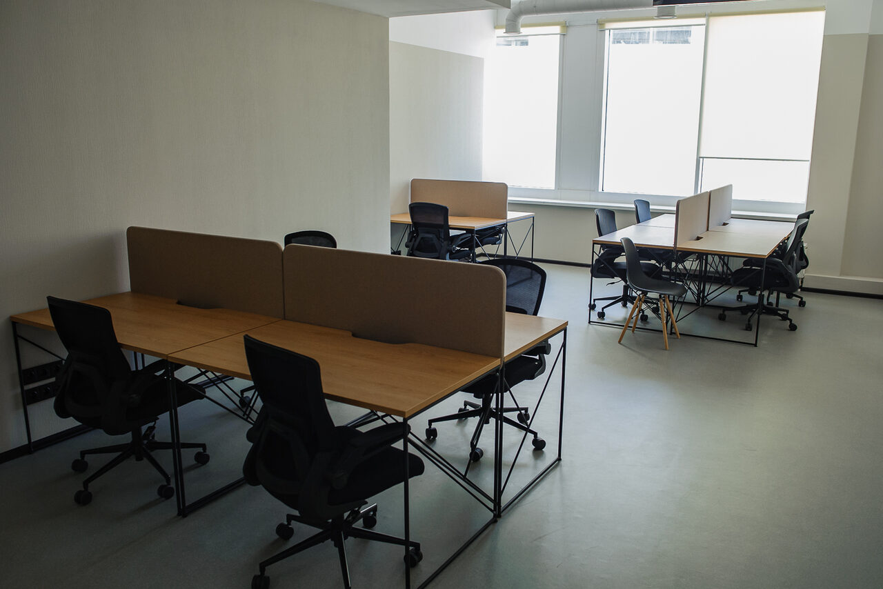 Арендовать Smart Coworking(Умный Коворкинг). Просторный офис с двумя группами столов. Каждая группа включает два стола с перегородками и черными офисными креслами. Светлые стены, большие окна, обеспечивающие естественное освещение. Пол светлый, потолок белый.
