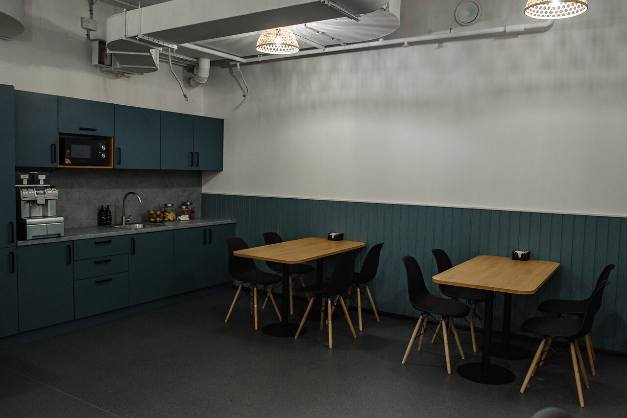 Арендовать Smart Coworking(Умный Коворкинг). Небольшая кухня с синими шкафами и серой столешницей. Имеется микроволновая печь, кофемашина и раковина. Две деревянные столы с черными стульями. Белые стены с синим панелированием. Потолок с системой вентиляции и подвесными светильниками.