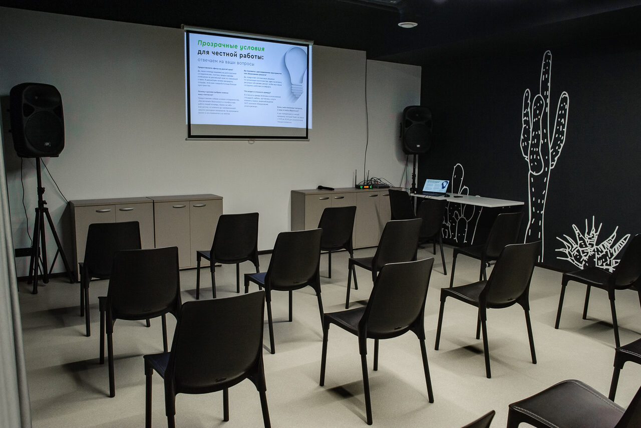 Арендовать Smart Coworking(Умный Коворкинг). Конференц-зал с рядами черных стульев, перед которыми размещены тумбы, на стене проектор с изображением, рядом колонки. Черные стены с белыми рисунками кактусов. Светлый пол, темный потолок, фрагмент стола с ноутбуком.