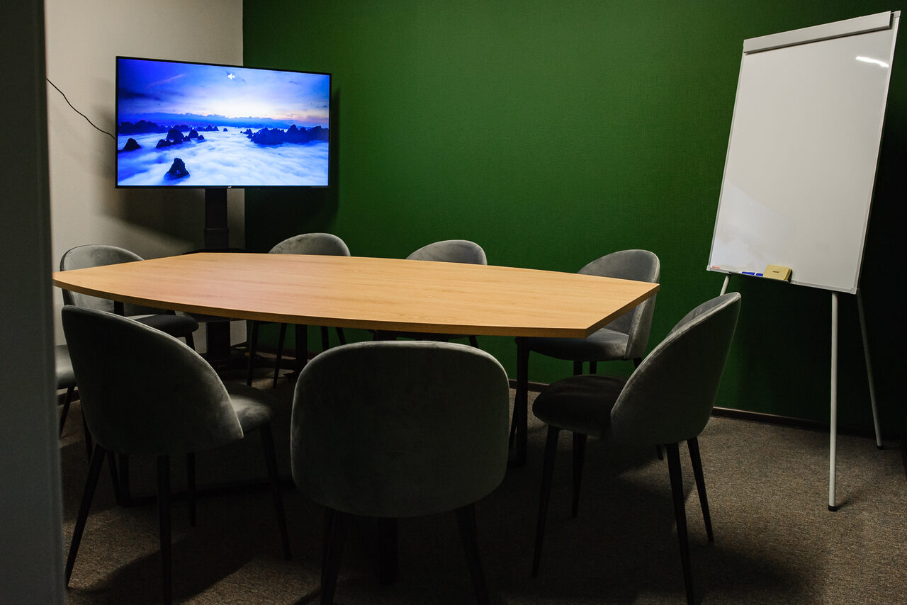 Арендовать Smart Coworking(Умный Коворкинг). Небольшое конференц-пространство с овальным деревянным столом, окружённым восемью серыми стульями. На стене смонтирован телевизор с природным изображением, рядом белая доска для записи. Зеленая стена, нейтральный пол.