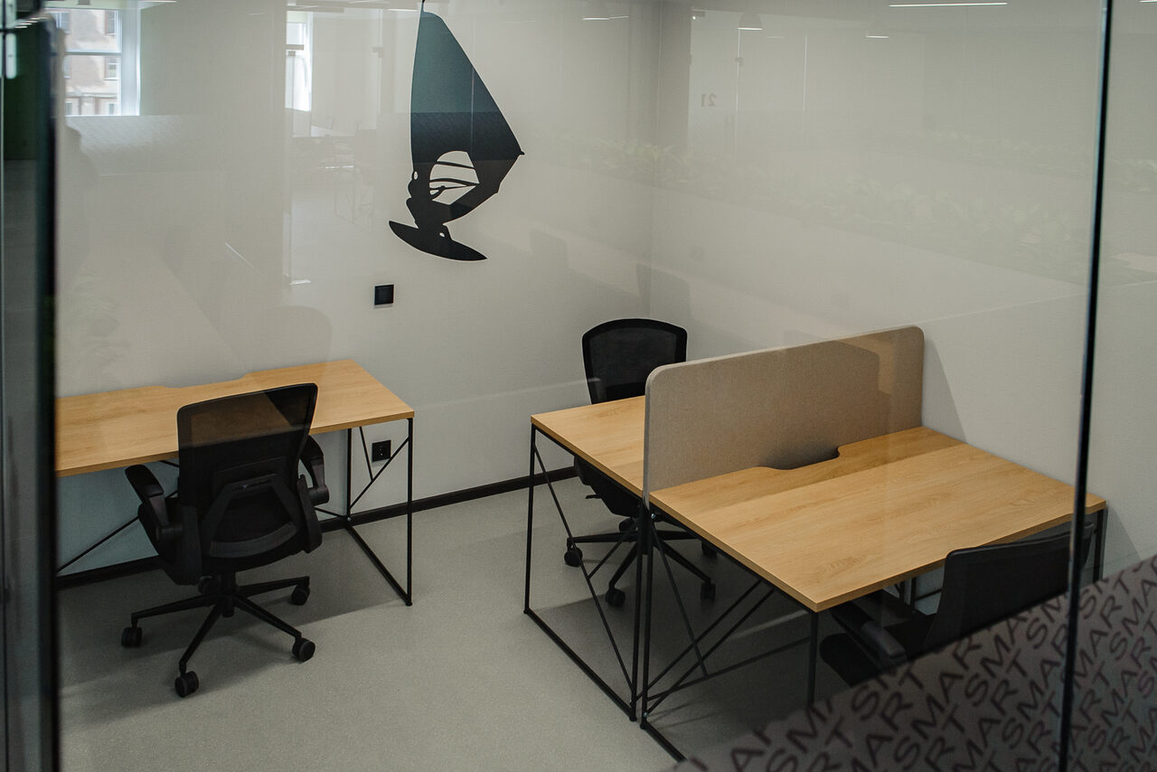 Арендовать Smart Coworking(Умный Коворкинг). Небольшой офис с двумя рабочими местами. У каждого рабочего места деревянный стол и черный офисный стул. На одном из столов установлена разделительная перегородка. Стены светлые, на одной - черный настенный декор. Пол светло-серый. Мягкое освещение.