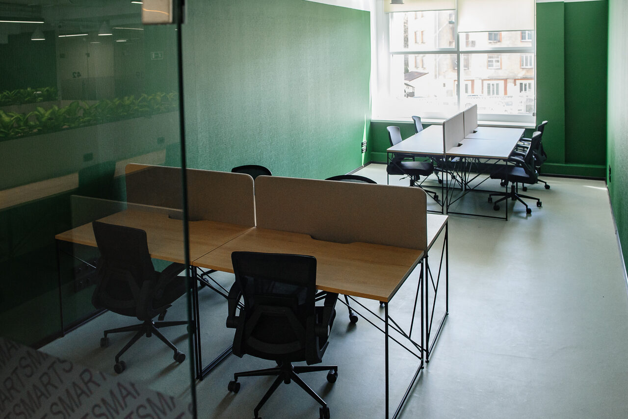 Арендовать Smart Coworking(Умный Коворкинг). Современный офис в зеленых тонах с перегородками. Пространство открытое, с рабочими столами и офисными стульями. Интерьер содержит стеклянные стены, окно, впускающее естественный свет. Стены зеленого цвета, полы светлые.
