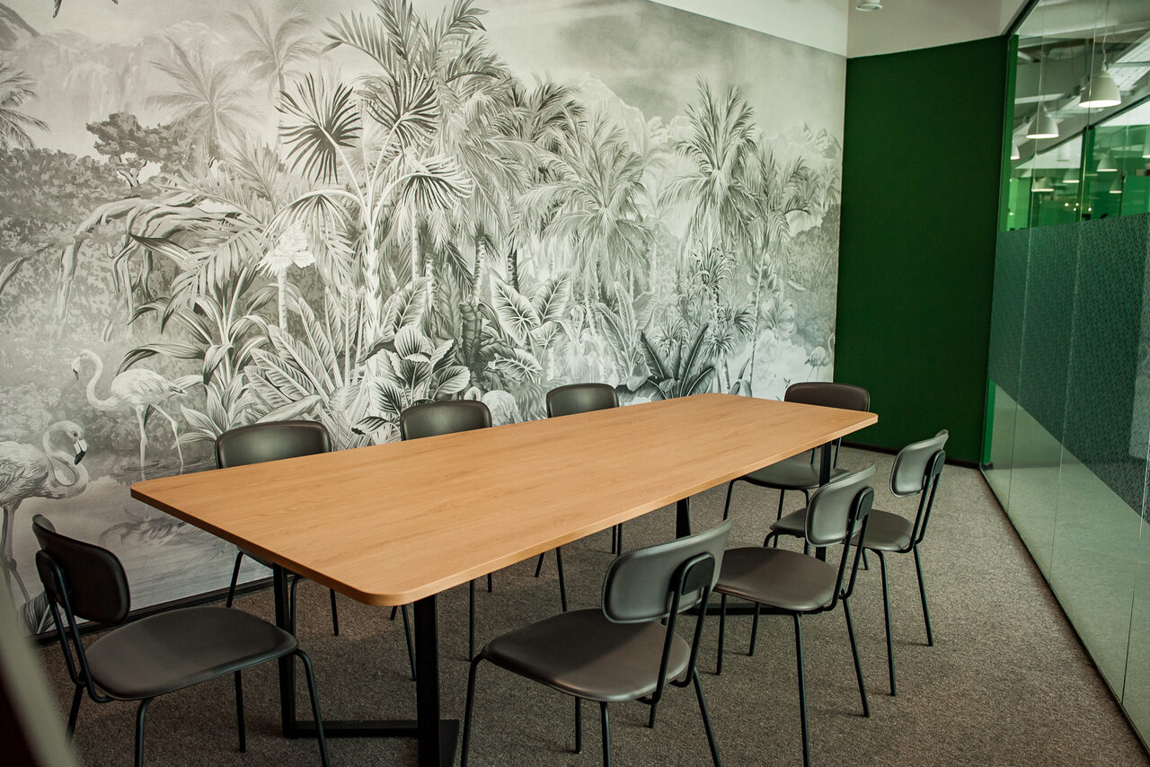 Арендовать Smart Coworking(Умный Коворкинг). Конференц-зал с длинным деревянным столом и восемью стульями. Стена украшена обоями с растительным узором. Пол ковровый, стеклянная перегородка, зеленая стеновая панель. Естественное освещение через большое окно.