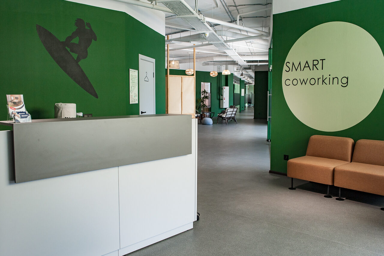 Арендовать Smart Coworking(Умный Коворкинг). Коворкинг-пространство с зелеными стенами, на переднем плане стойка ресепшн серо-белого цвета, рядом коричневый диван. На стене круглая эмблема с текстом "SMART coworking". В глубине видны столы и стулья.