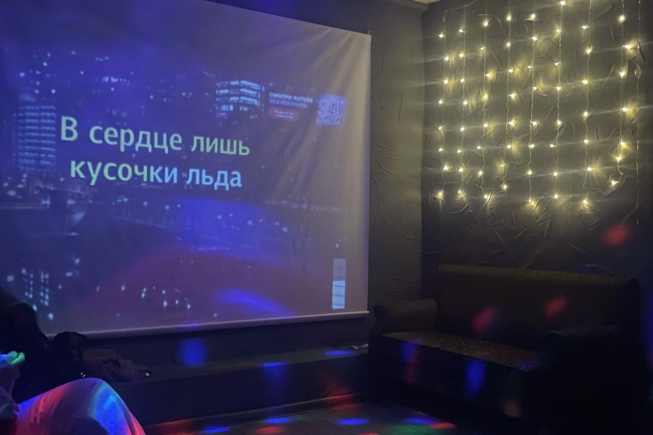 Арендовать Лофт ВЕЧЕ’Р. Тёмная комната с проектором, текст на экране "В сердце лишь кусочки льда". Справа тёмный диван. На стене светодиодные гирлянды. Локальное освещение создает разноцветные пятна на полу. Стены тёмные, текстурированные.