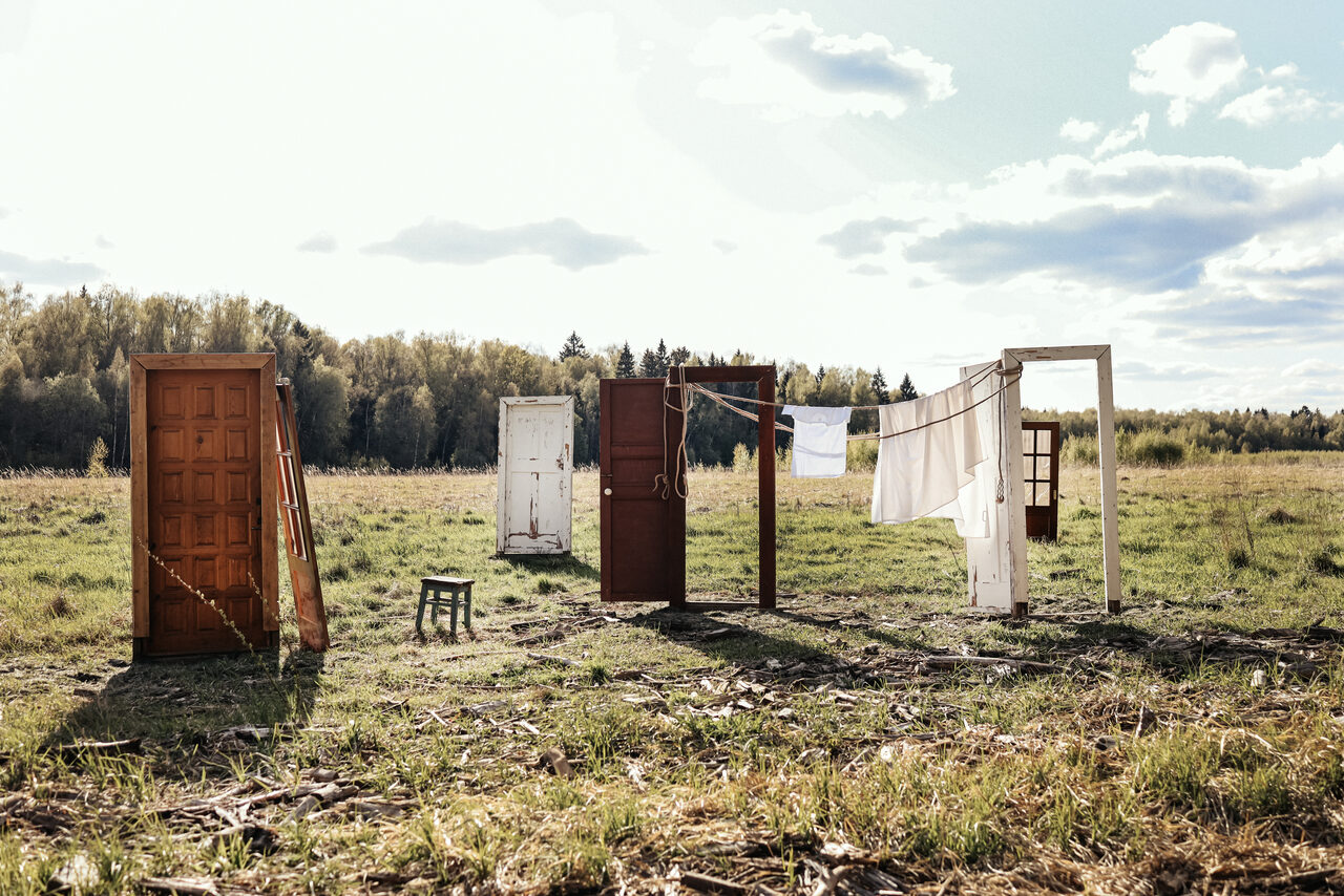 Арендовать Фотостудия Воздух. Поле с зелёной травой, несколько дверей и рам, раскладной стул, верёвка с развешенным бельём, солнечный день, деревья на горизонте, облака в небе.