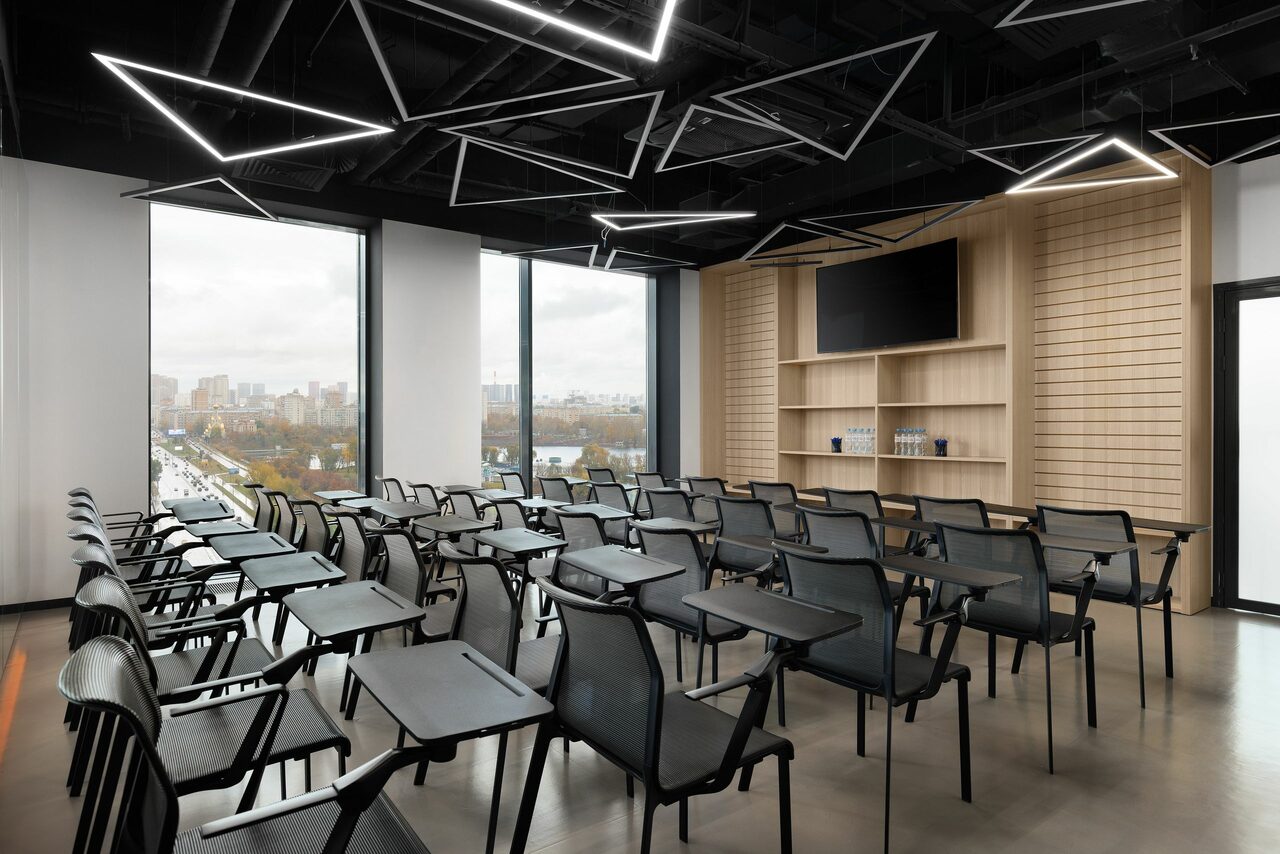 Арендовать YES Technopark(ДА Технопарк). Конференц-зал. Современное офисное пространство с большим количеством стульев и столиков для конференций. Черные потолочные светильники геометрической формы, светлые деревянные панели на стене с экраном, большие окна с видом на город.