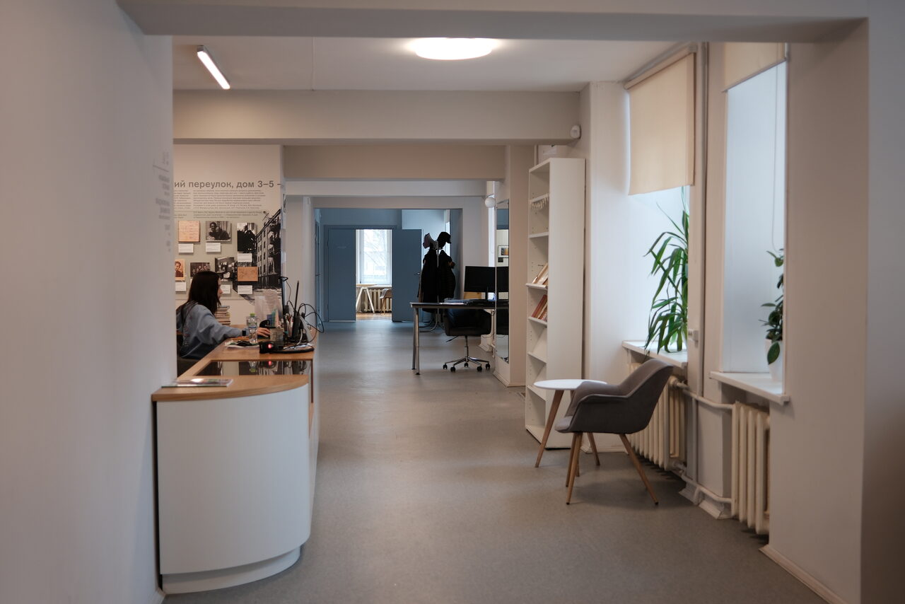 Арендовать Лекторий Научно-просветительского центра Музея М.А. Булгакова. Просторный офис с серыми стенами и полом, большая стойка ресепшн с компьютером слева, пара кресел и стеллаж с книгами справа. В глубине комнаты стол с компьютером, окна с жалюзи, несколько растений. Освещение потолочное.