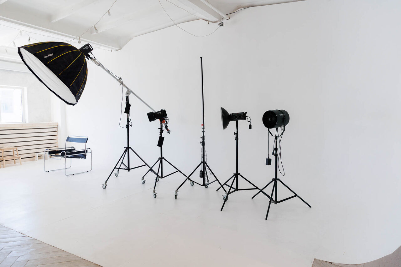 Арендовать Art Room Studio (Арт Рум). Фотостудия. Студийное пространство для фотосъёмки, белые стены и пол, профессиональное осветительное оборудование на стойках, светодиодные лампы, оконный проем с естественным светом, небольшая скамья, стул с металлическим каркасом, светлая древесина на полу.