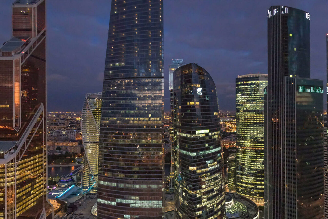 Арендовать Офис с панорамным видом. Высотные стеклянные здания в городской застройке, множество офисных небоскрёбов с вечерним внутренним освещением, тёмное небо, панорамный вид на город с огнями улиц, вечернее время, современные архитектурные формы.