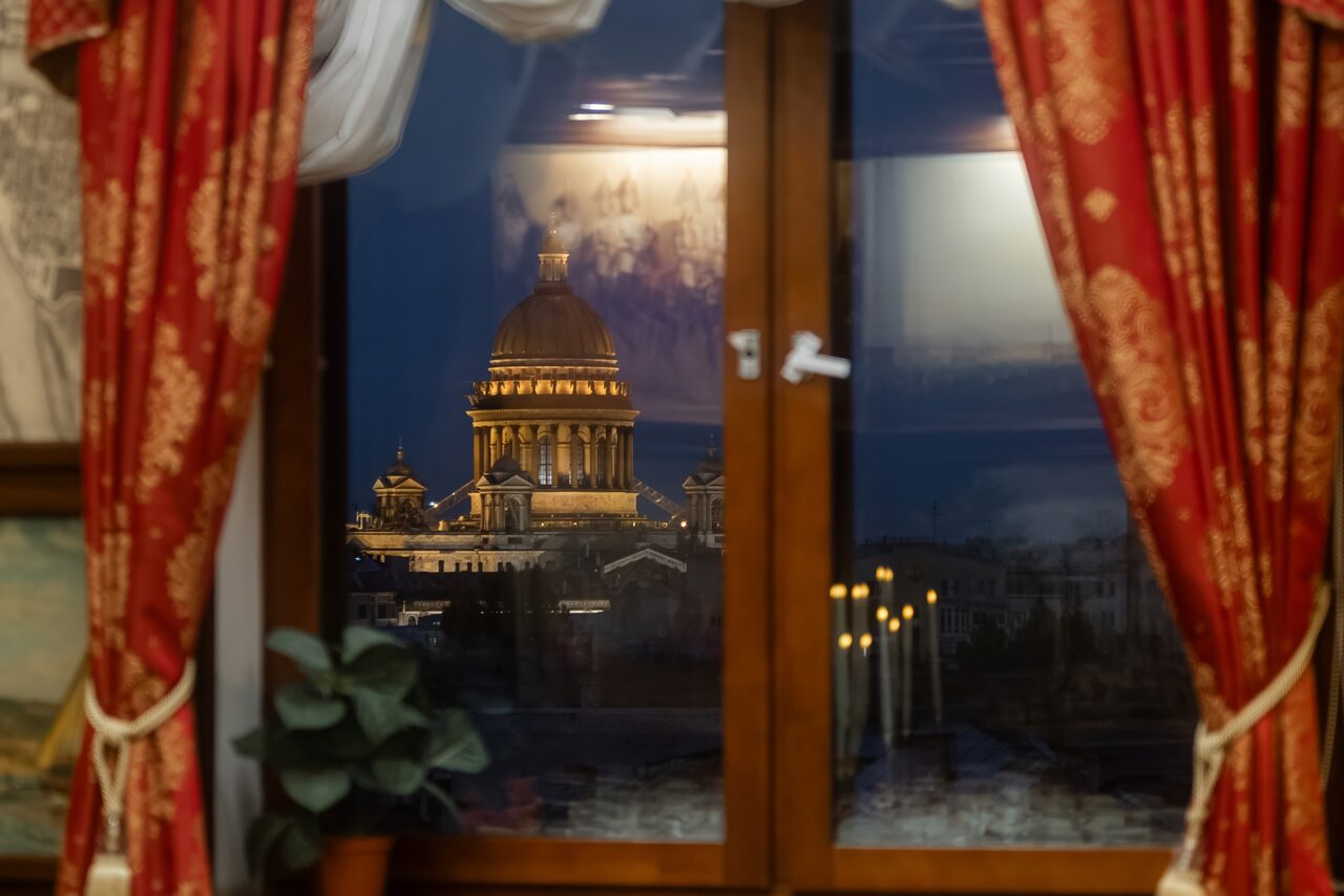 Арендовать Амбассадор Отель. Окно с видом на освещённое здание с куполом в ночное время. Деревянная оконная рама, красные занавески с узорами, комнатное растение в горшке на подоконнике.