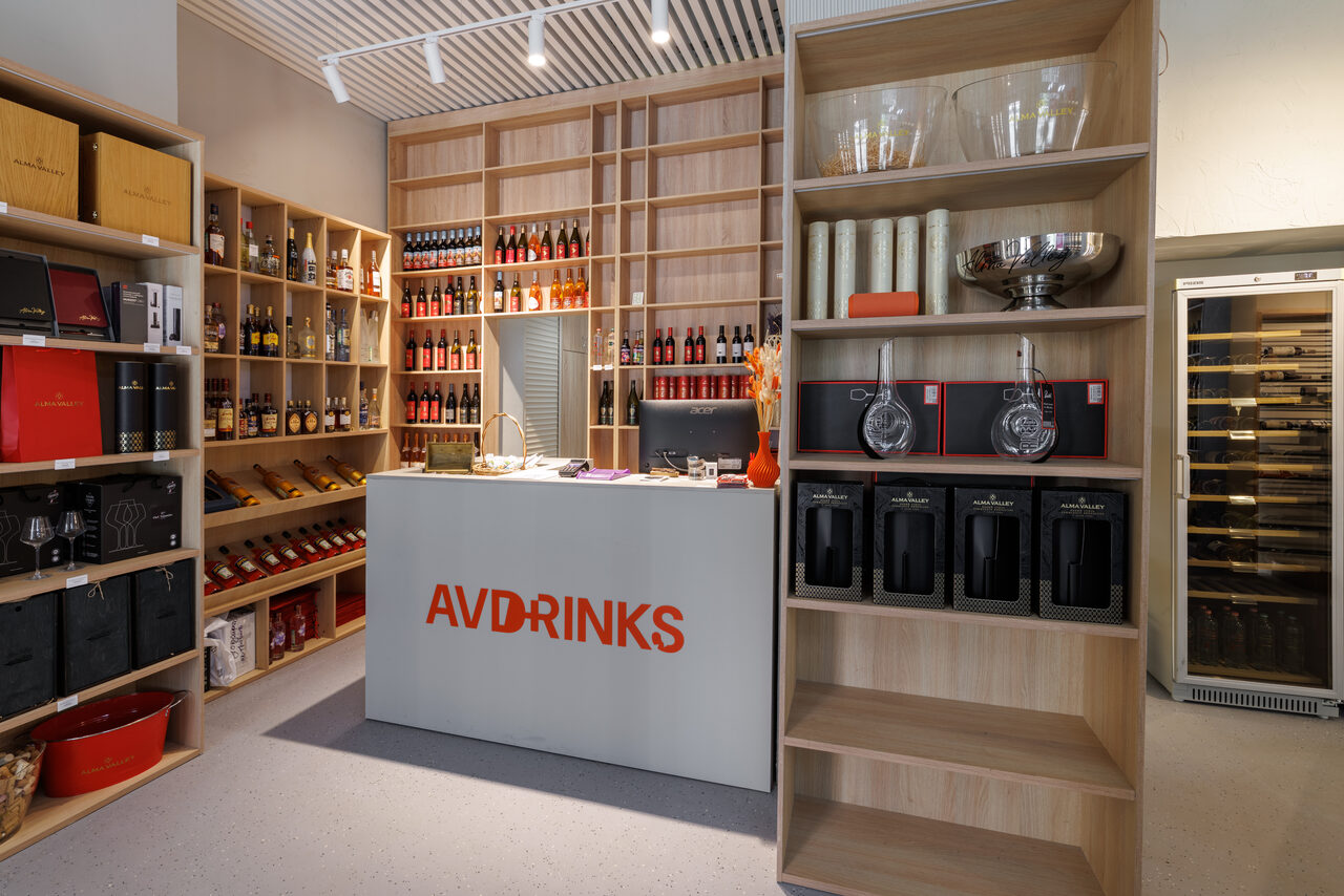 Арендовать Винотека AVDRINKS(АВДРИНКС) на 12 гостей. Пространство магазина; деревянные стеллажи с напитками и аксессуарами; стол с надписью AVD на передней панели; подсветка трековыми светильниками; полки заполнены товарами; нейтральные цвета, современный интерьер.