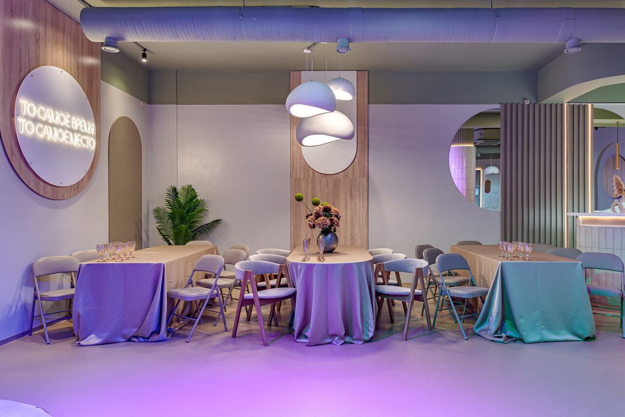 Арендовать SOK SPACE (Сок Пространство). Лофт Октябрь. Современное пространство с круглым и прямоугольными столами, покрытыми скатертями в мягких тонах. Складные стулья вокруг столов. Декор: растения, вазы с цветами. На стене неоновая надпись. Потолочные светильники, нейтральные стены, круглые зеркала.