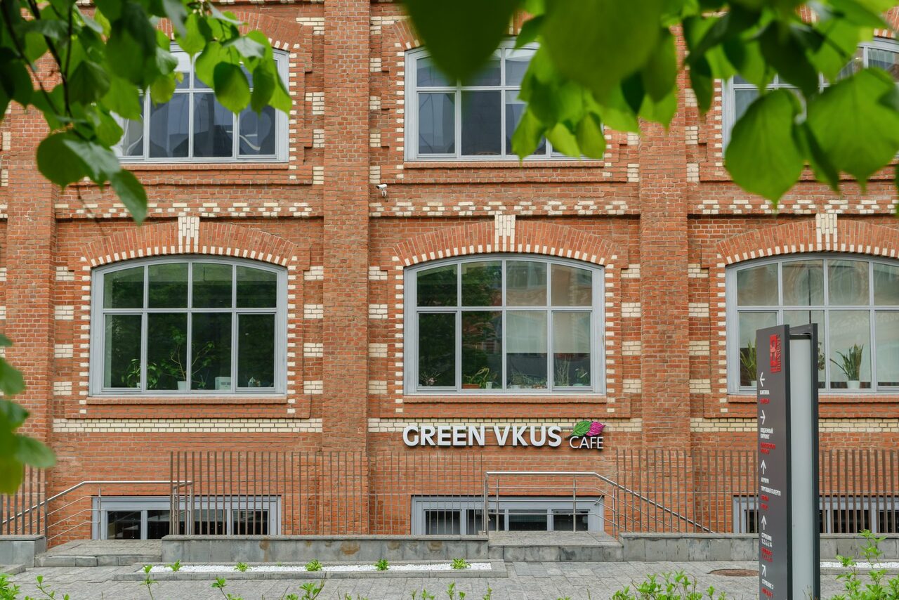 Арендовать Green Loft(Зеленый Лофт). Красное кирпичное здание с большими арочными окнами. Вывеска со словами "GREEN VKUS CAFE" на фасаде. Перед зданием металлографитовый забор и каменные ступени. Деревья на переднем плане.