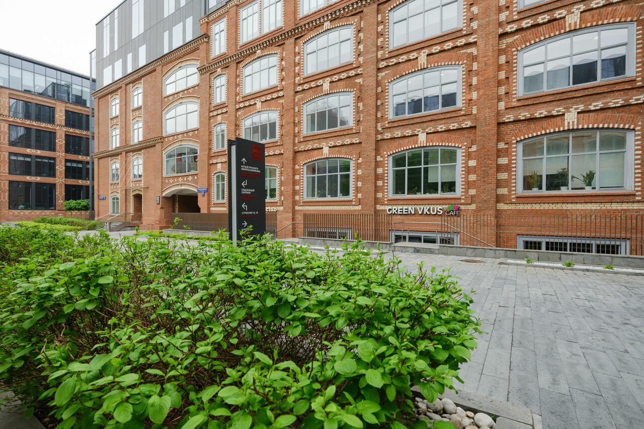 Арендовать Green Loft(Зеленый Лофт). Красное кирпичное здание с большими арочными окнами. На переднем плане тротуар из серых плит и кусты. Видно черную стойку с текстом. Вывеска "GREEN VKUS" на фасаде здания. Обстановка ухожена, пешеходная зона.