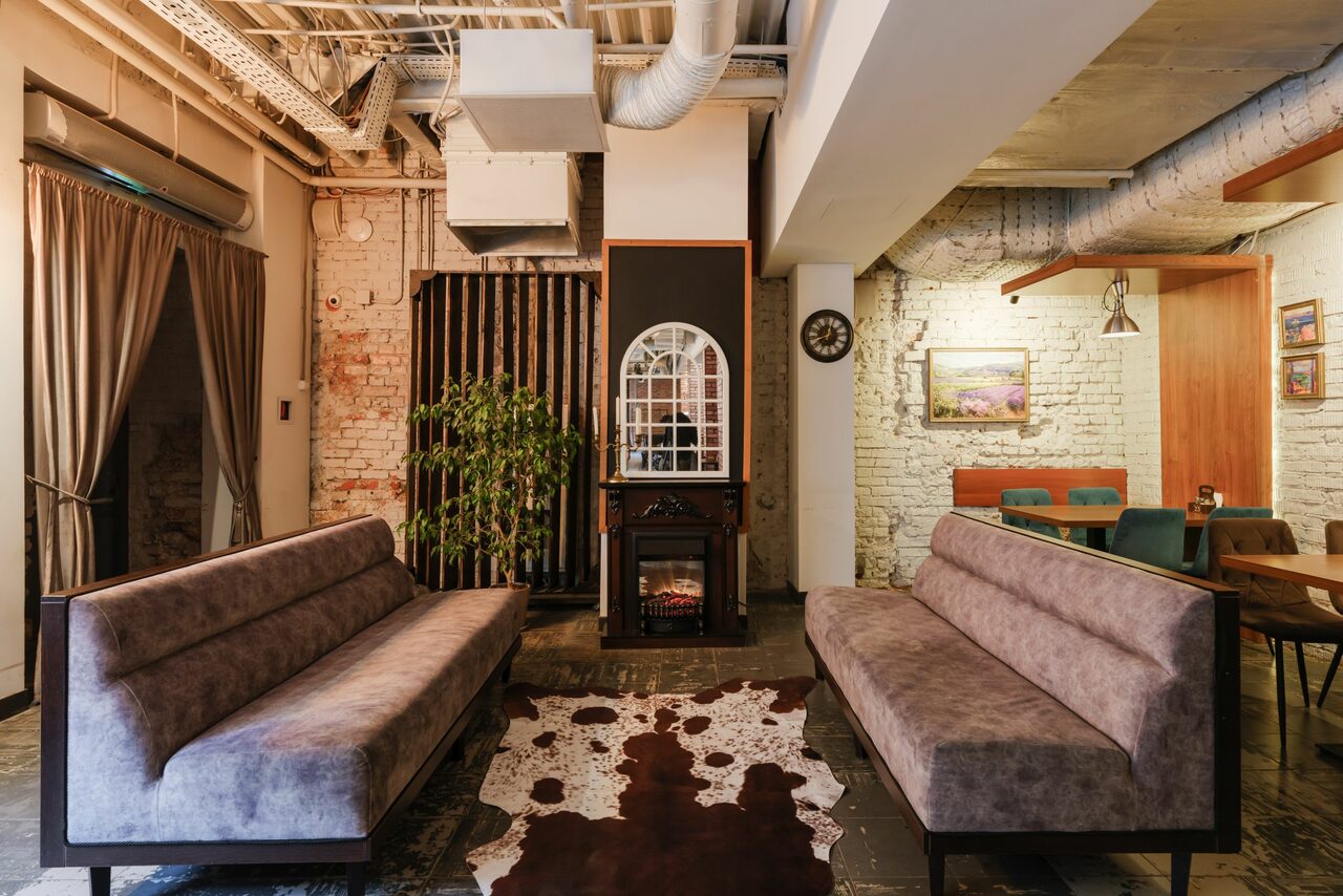 Арендовать Green Loft(Зеленый Лофт). Лофт-пространство с открытыми коммуникациями на потолке, кирпичные стены, два серых дивана, камин, зеркало, ковёр с черно-белым узором, деревянный стол с зелеными стульями. Освещение - подвесные лампы. Стены белого и кирпичного цвета. Пластиковое растение.